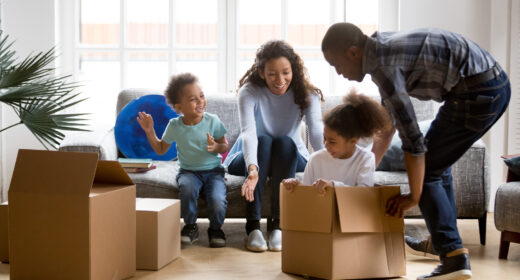 Une grande famille afro-américaine joyeuse et enjouée emménage dans un nouvel appartement ; la petite fille d'âge préscolaire est assise dans des cartons, le père la fait rouler vers la mère, ils jouent ensemble ; concept d'achat immobilier
